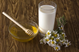 A glass of milk with chamomile flowers and honey next to it on a table.