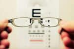 A pair of hands holding eye glasses in front of a Snellen chart.